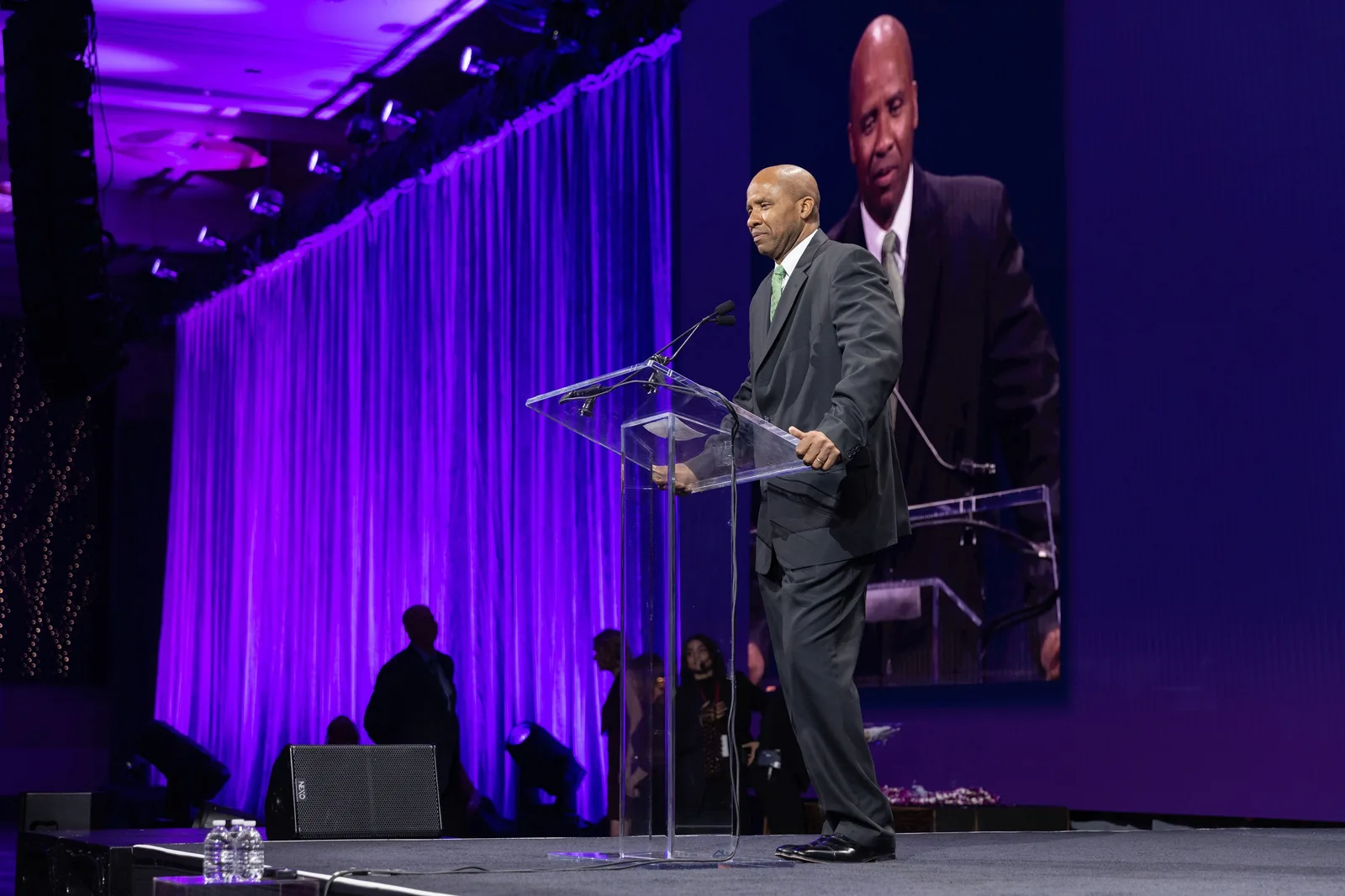 Rutherford Pascal speaking at a glass lectern, his face on the screen behind him